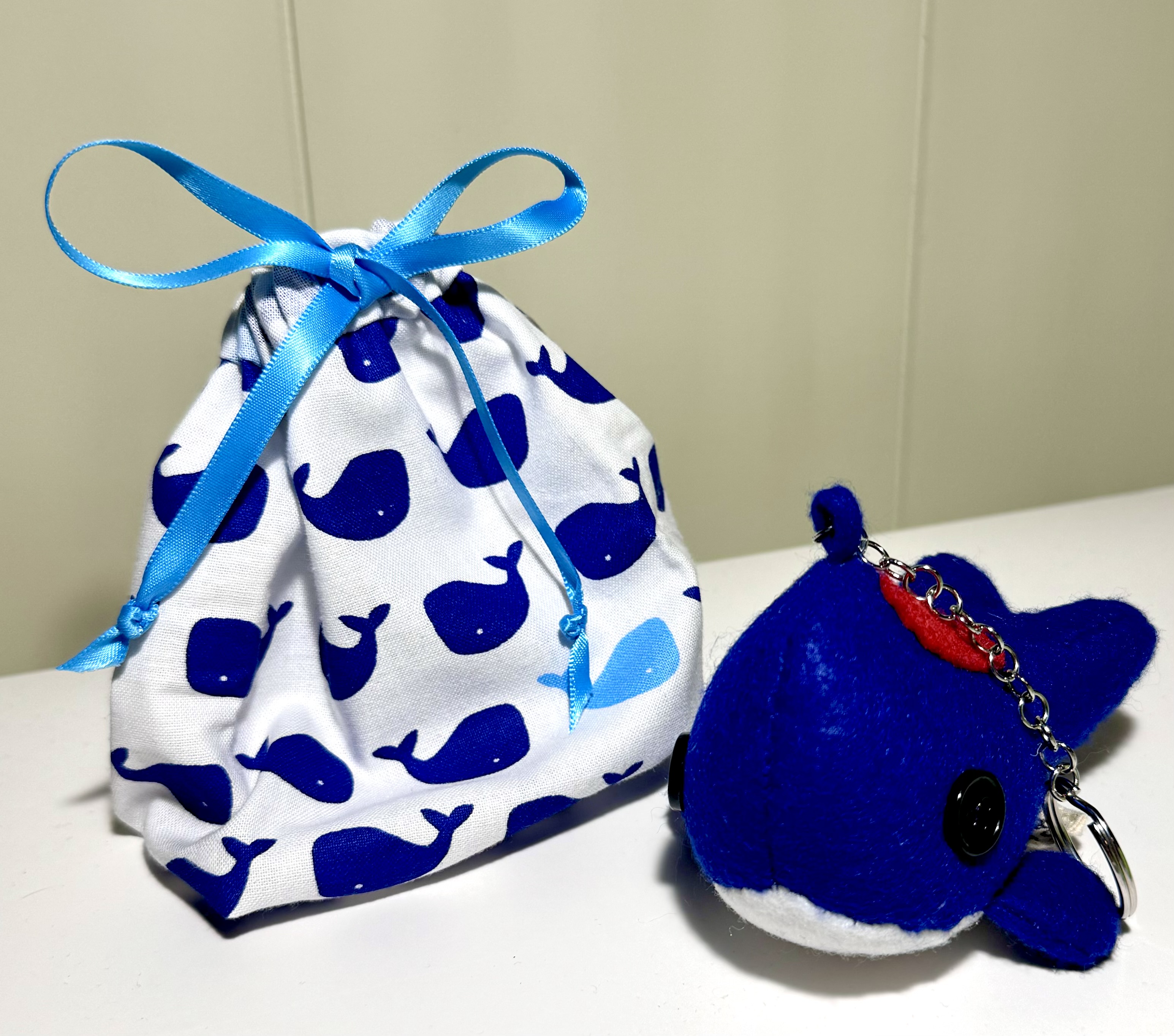 A keychain style red patch Heart Whale on it's back sits in front of a drawstring bag made from a whale print cloth